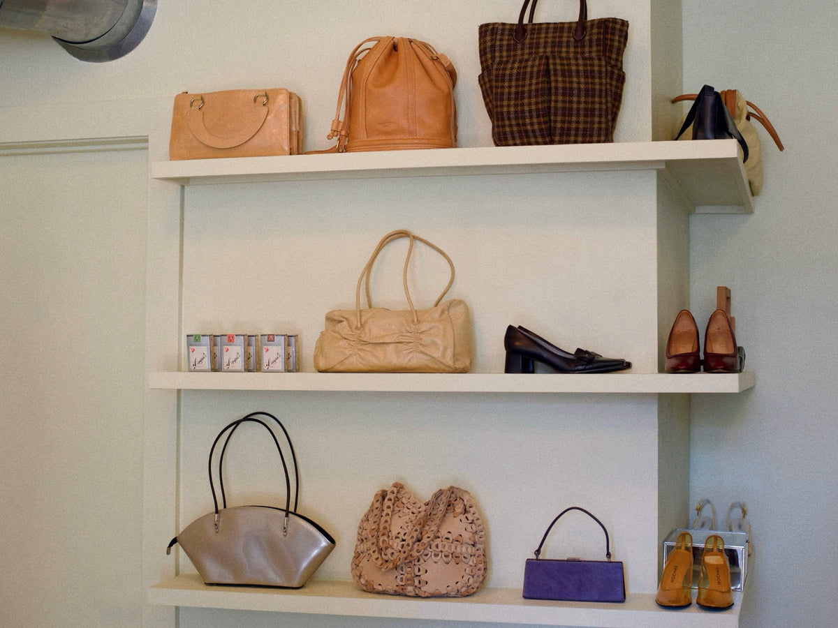 A set of three white floating shelves displaying an assortment of handbags, shoes, and accessories against an off-white wall.